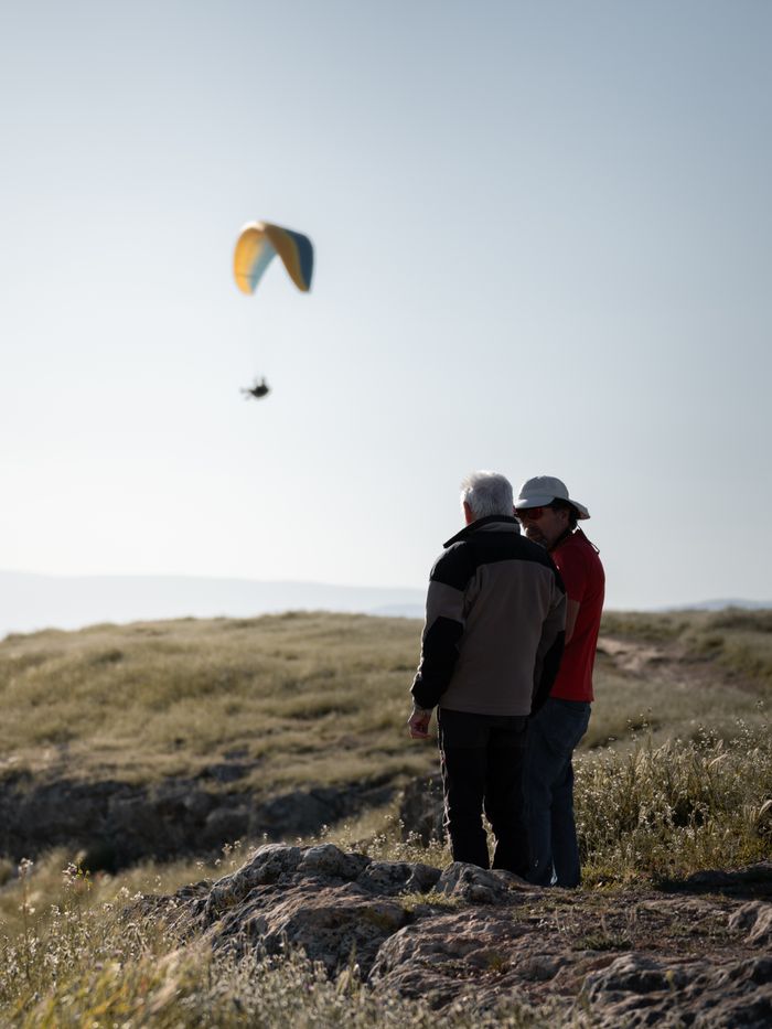 Two men in rural setting watching a paraglider in the sky.